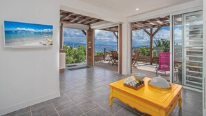 A spacious room with a large window offering a view of the beach and ocean. There is a yellow table with a chessboard and a bowl.