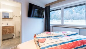 Een slaapkamer met een bed, een aan de muur bevestigde televisie, een raam met uitzicht op een besneeerd landschap en een badkamer met een wastafel en spiegel.