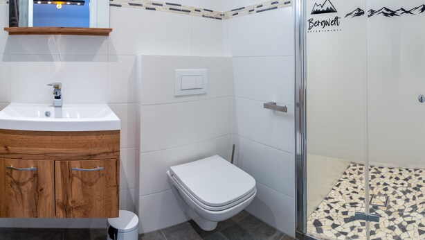 Une salle de bain avec un meuble-lavabo en bois, des toilettes blanches, une douche avec une porte en verre et un sol carrelé.