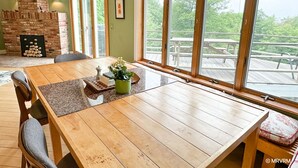 Un comedor con mesa de madera, sillas, chimenea de ladrillo y ventanas amplias con vista a una terraza.