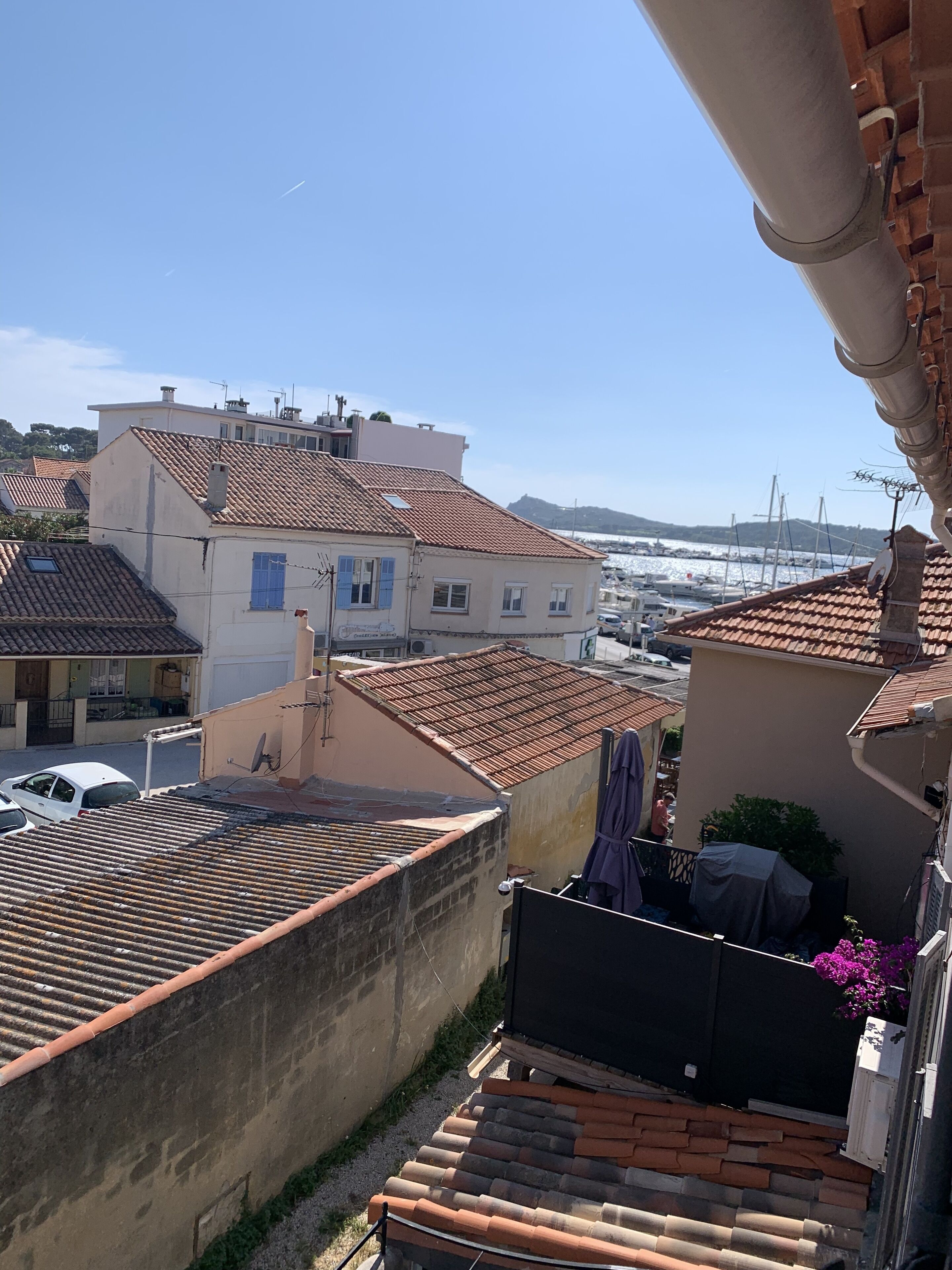 Zona residenziale con tetti in tegole di terracotta, un edificio bianco con persiane blu e vista su un porto turistico con barche.
