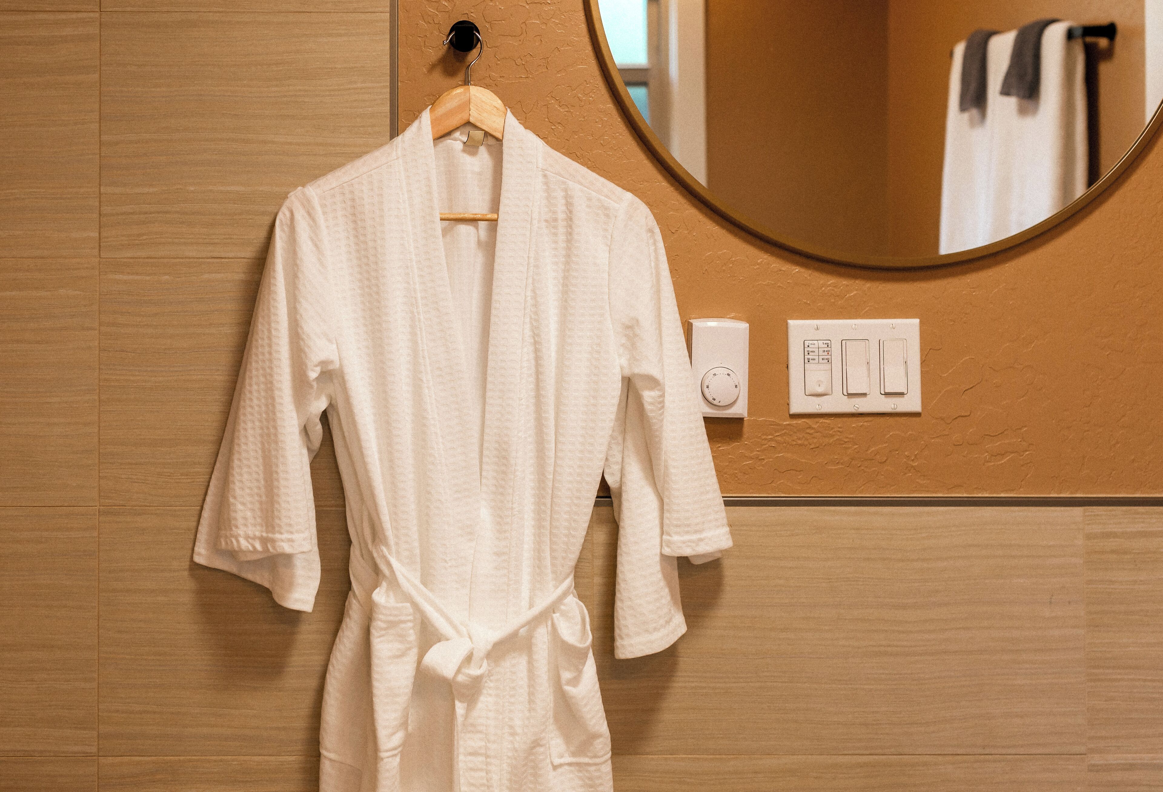 A white bathrobe hanging on a wooden hanger in a bathroom with a round mirror and a thermostat on the wall.