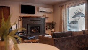 A living room with a fireplace, a sofa, a television, and a window with blinds. - Escape to this artsy 2-bedroom MCM Guesthouse in charming Phoenix, OR. Unwind in the luxurious spa soaking tub after exploring Southern Oregon. Features high-end design for a stylish, quiet retreat. (Phoenix)
