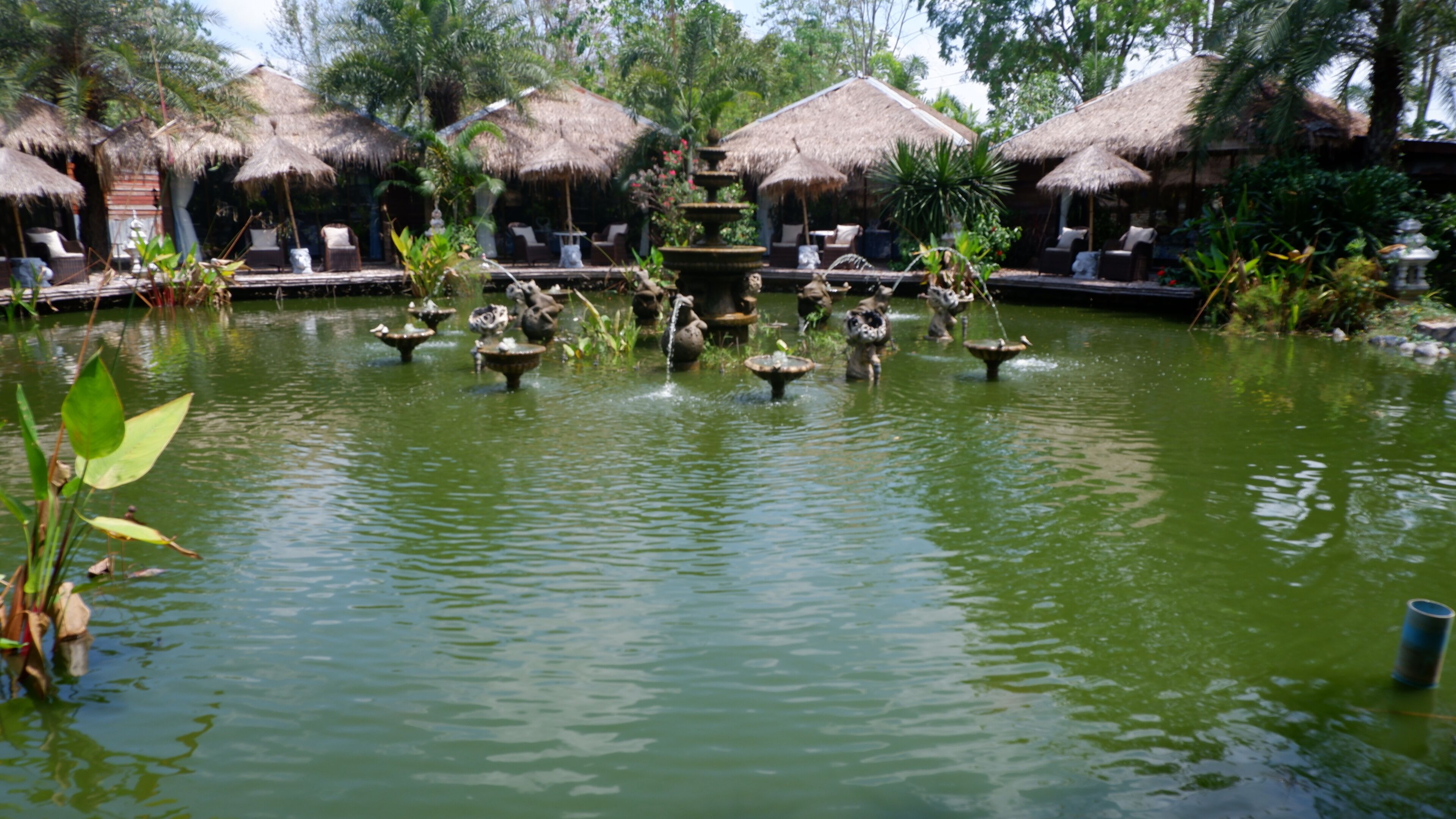 A tranquil pond with multiple fountains surrounded by thatched-roof huts and lounge chairs.