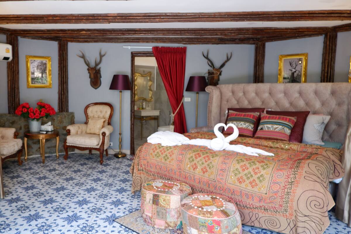 A bedroom with a large bed, a chair, a small table, and a mirror. There are deer antlers on the wall and a red curtain.