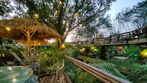 Estructura de techo de paja con un puente de madera sobre un elemento acuático, rodeada de exuberante vegetación.