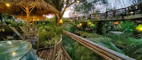 A thatched-roof structure with a wooden bridge over a water feature, surrounded by lush greenery.