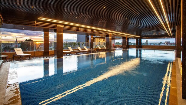 An indoor swimming pool with lounge chairs and a sunset view through large windows. - Horison Diana Timika (Timika)