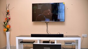 A flat-screen TV mounted on a wall above a white entertainment center with a glass shelf and a black drawer. - Stunning 2-bed Apartment in Namugongo Town Kampala (Kampala)