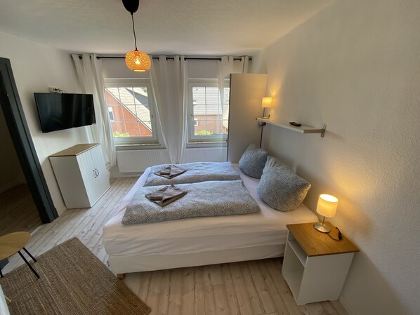 Une chambre à coucher avec un lit, une table de chevet, une lampe, une télévision, des fenêtres et des rideaux.