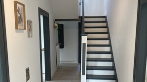 Un pasillo con piso de madera, una escalera con escalones negros y una puerta que da a otra habitación.