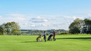 Un campo de golf con cielo despejado, árboles y una colina distante.