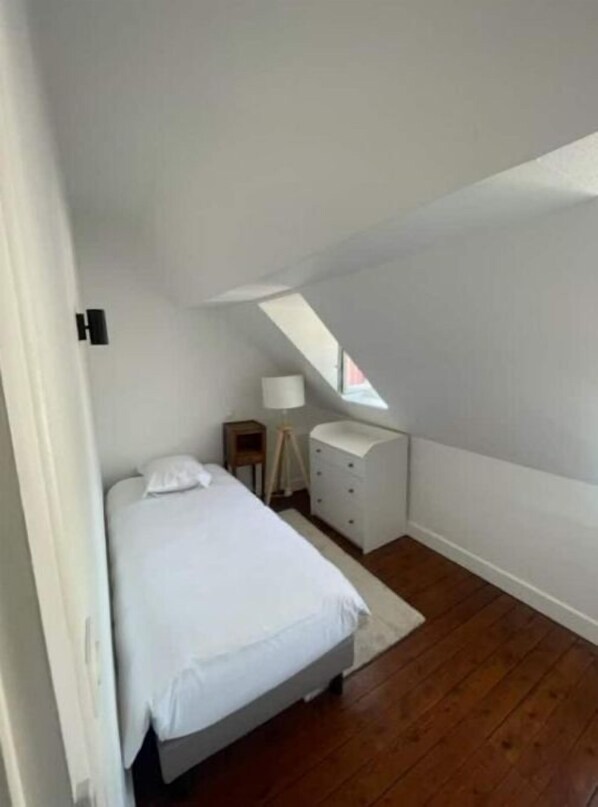 A small, white, single-bed room with a wooden floor, a bedside table, a lamp, and a small dresser. - Le Clos de Russy (Aure sur Mer)
