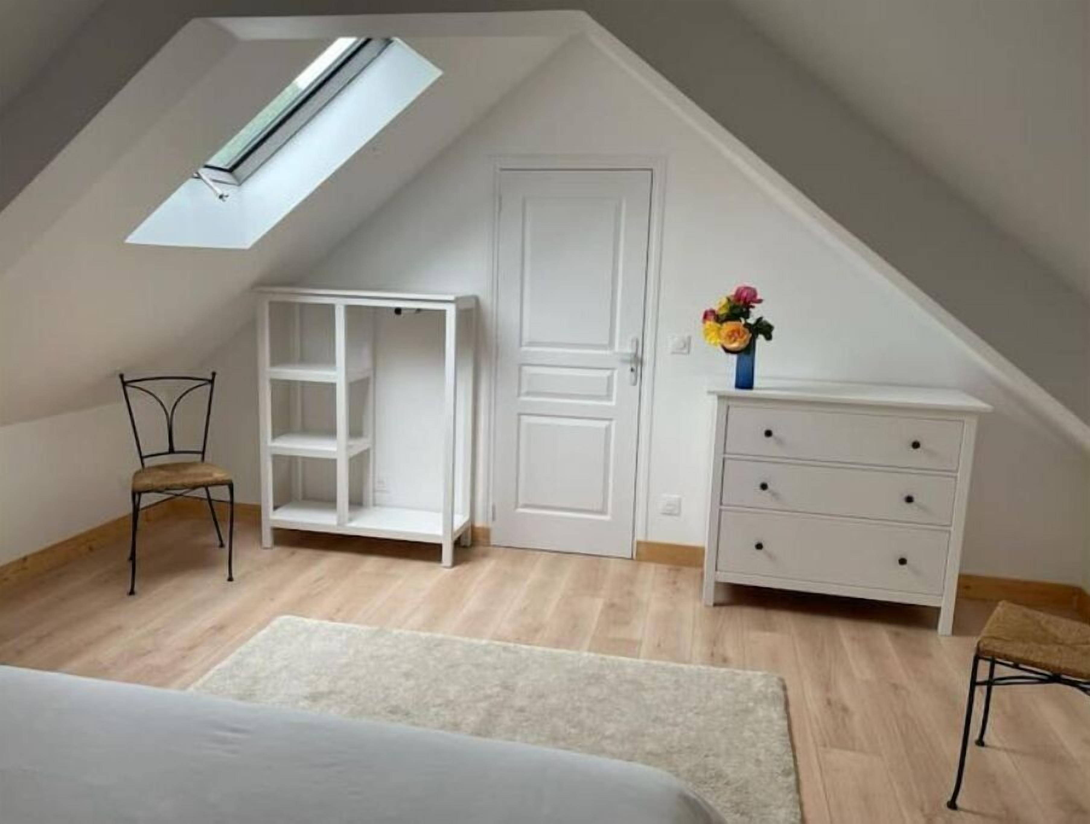 A room with a sloped ceiling, a skylight, a white wardrobe, a chair, and a small table with a vase of flowers.