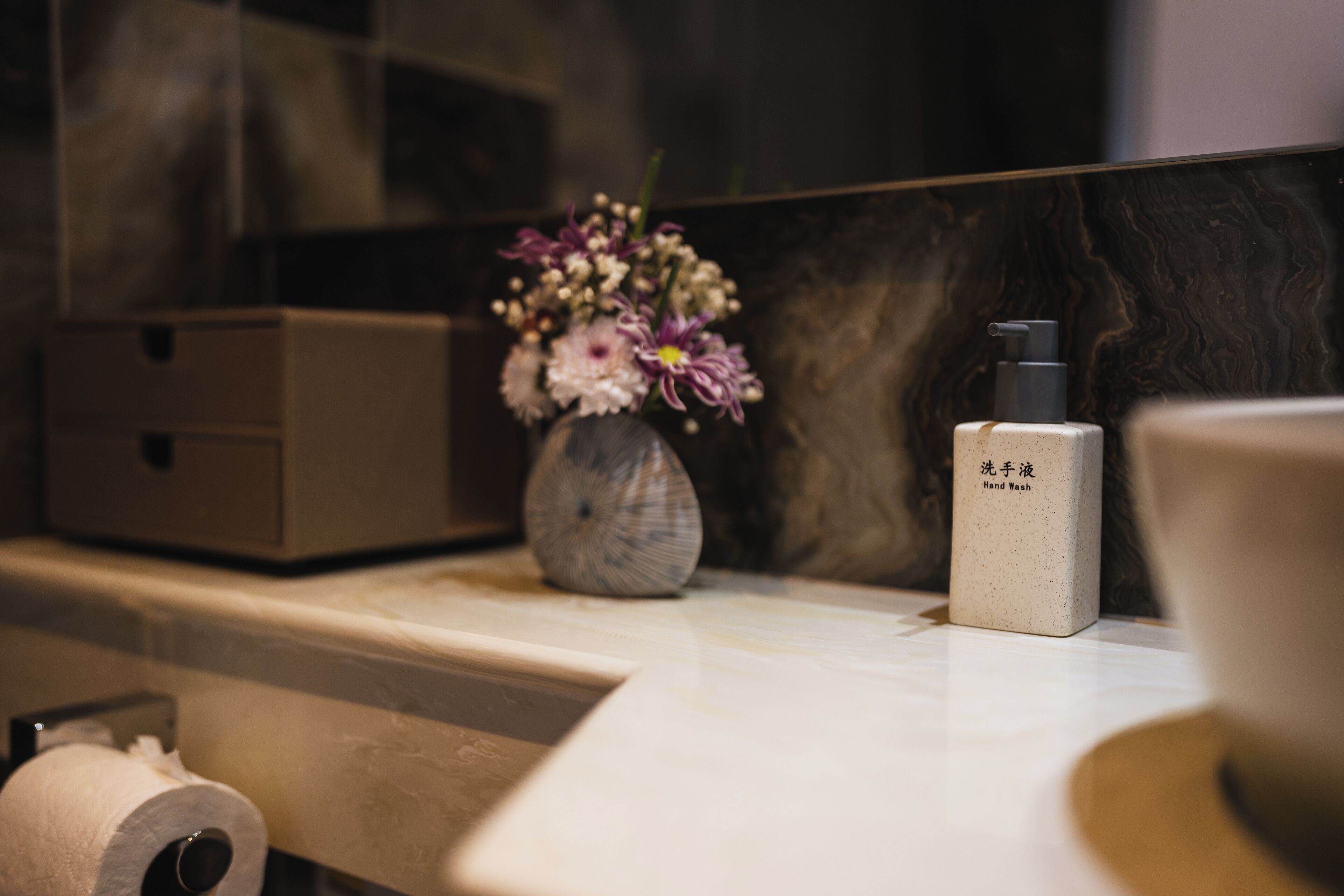 A bathroom vanity with a marble countertop, a vase of flowers, a hand soap dispenser, and a toilet paper roll.