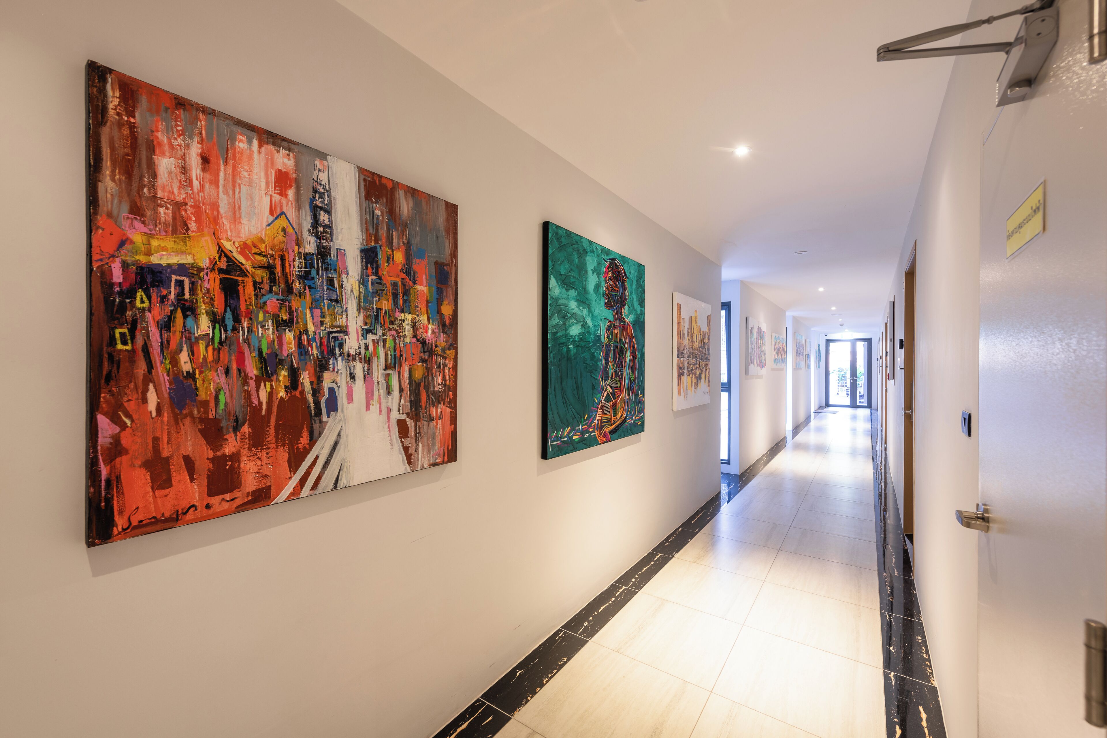 A hallway with two large abstract paintings on the walls, a tiled floor, and a door on the right.