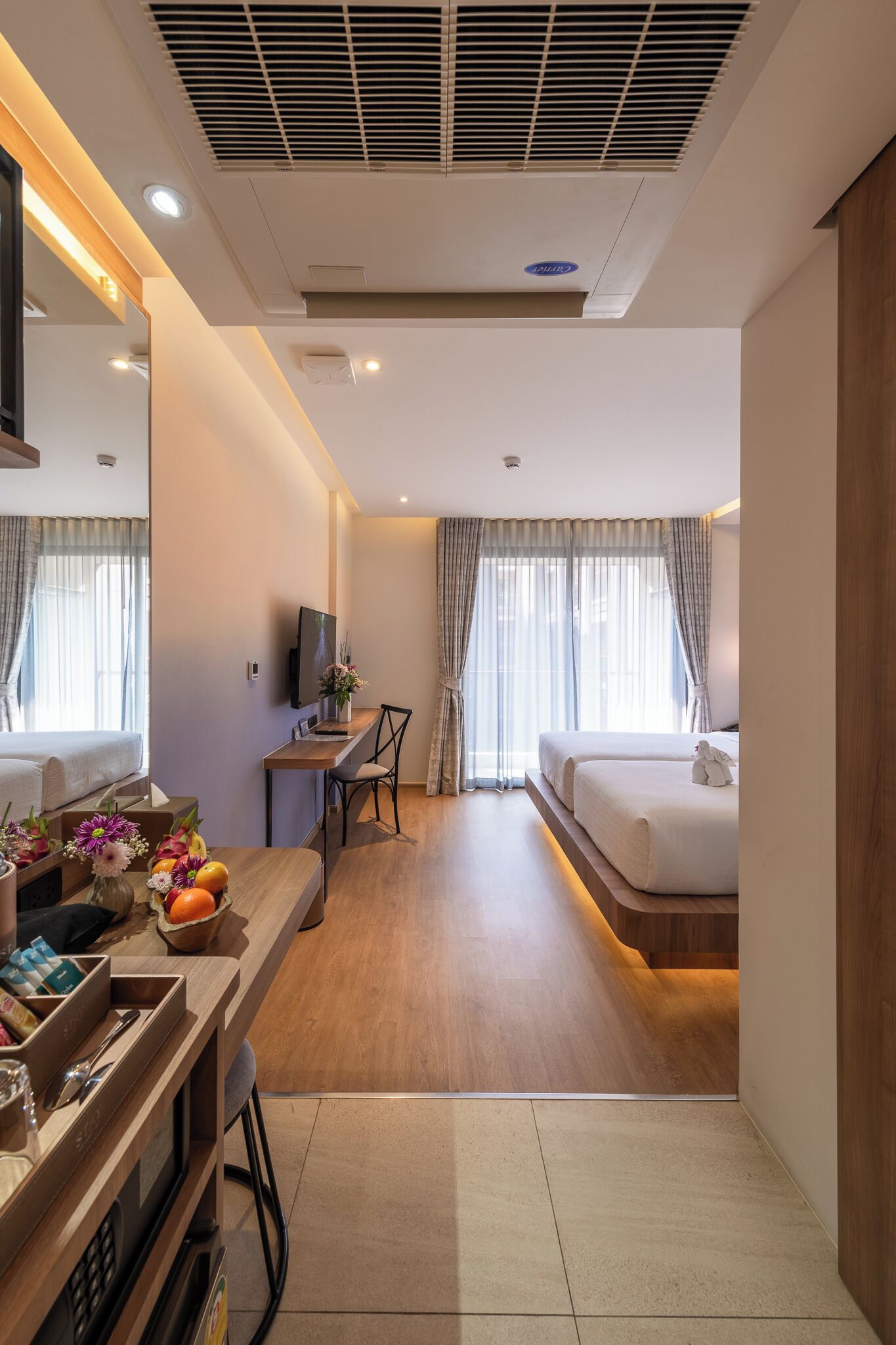 A hotel room with a bed, a desk, a chair, a TV, and a small table with flowers and fruits.
