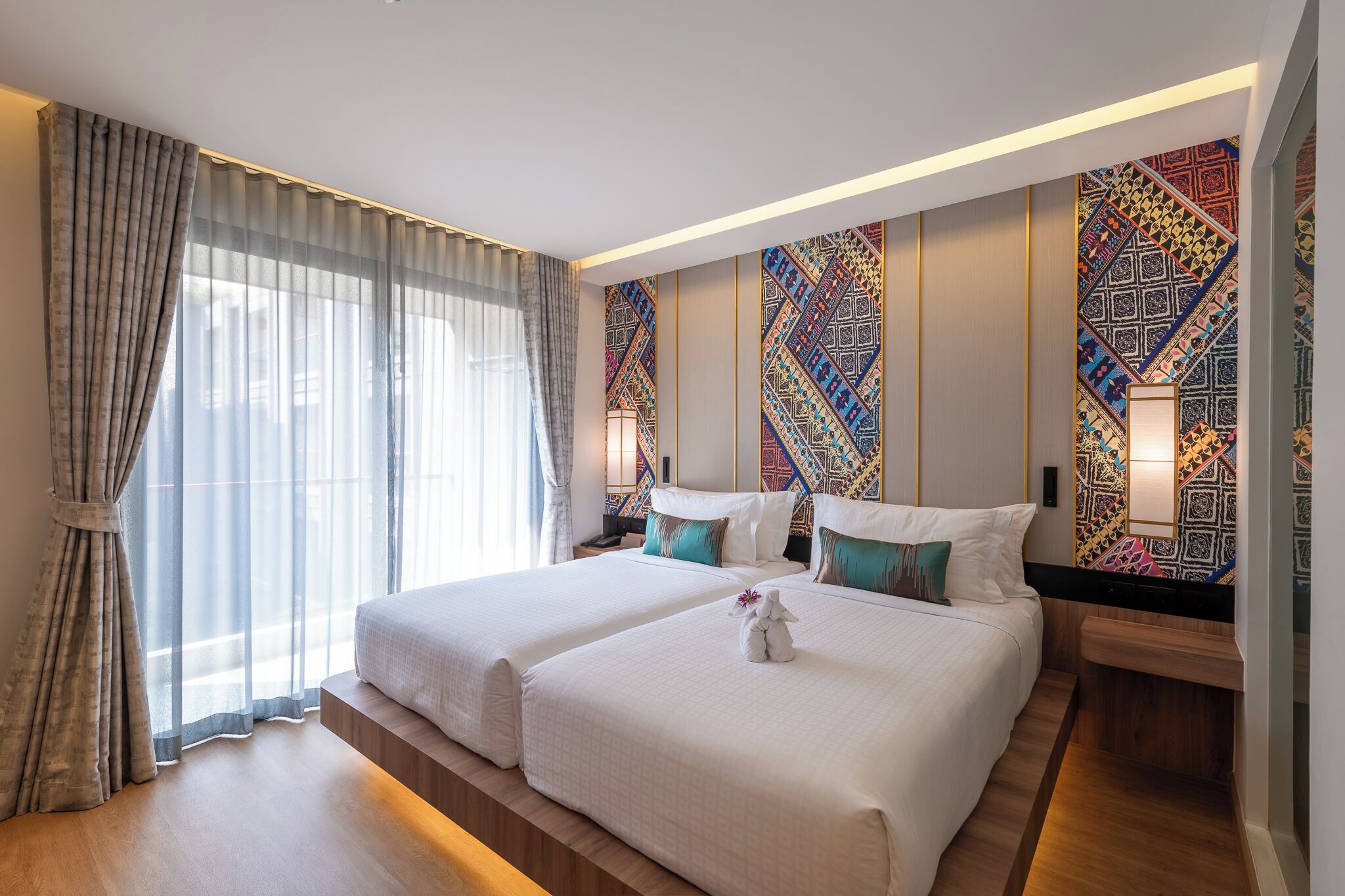 A hotel room with two beds, a large window with curtains, a patterned wall, and a nightstand with a lamp.