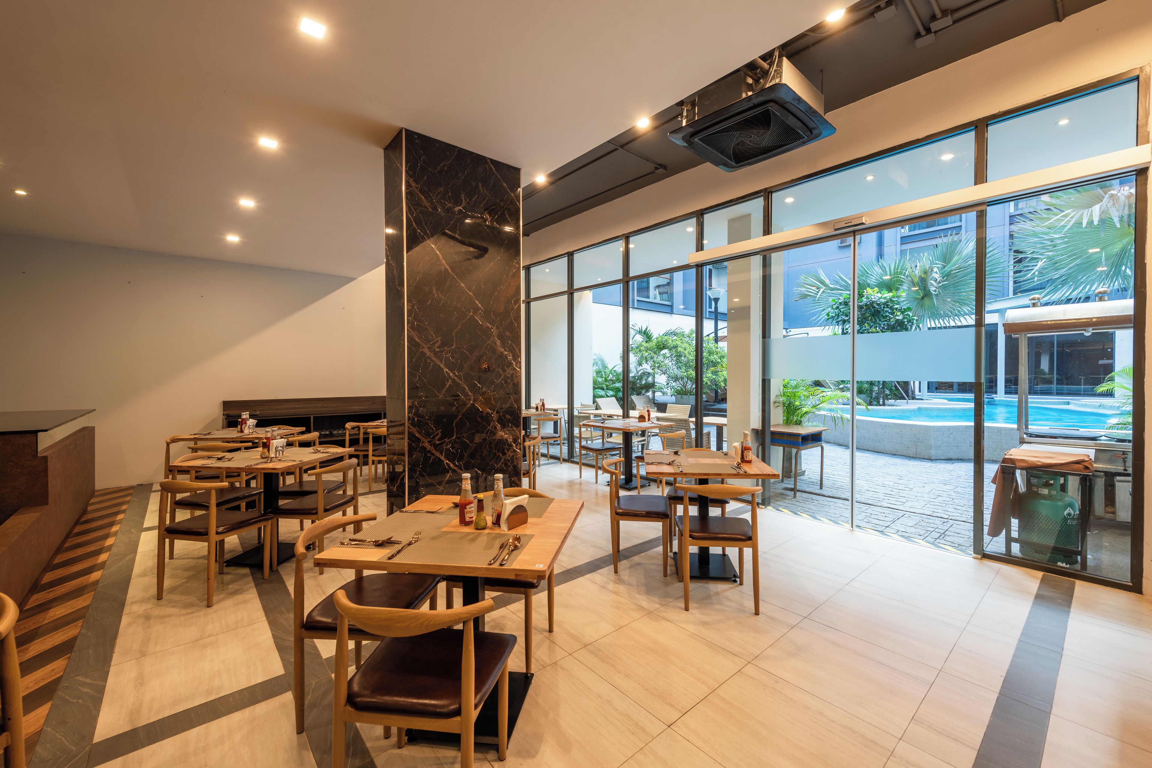 En modern restaurang med träbord och stolar, en marmorkolonn och stora fönster som ger utsikt över utomhuspoolområdet.