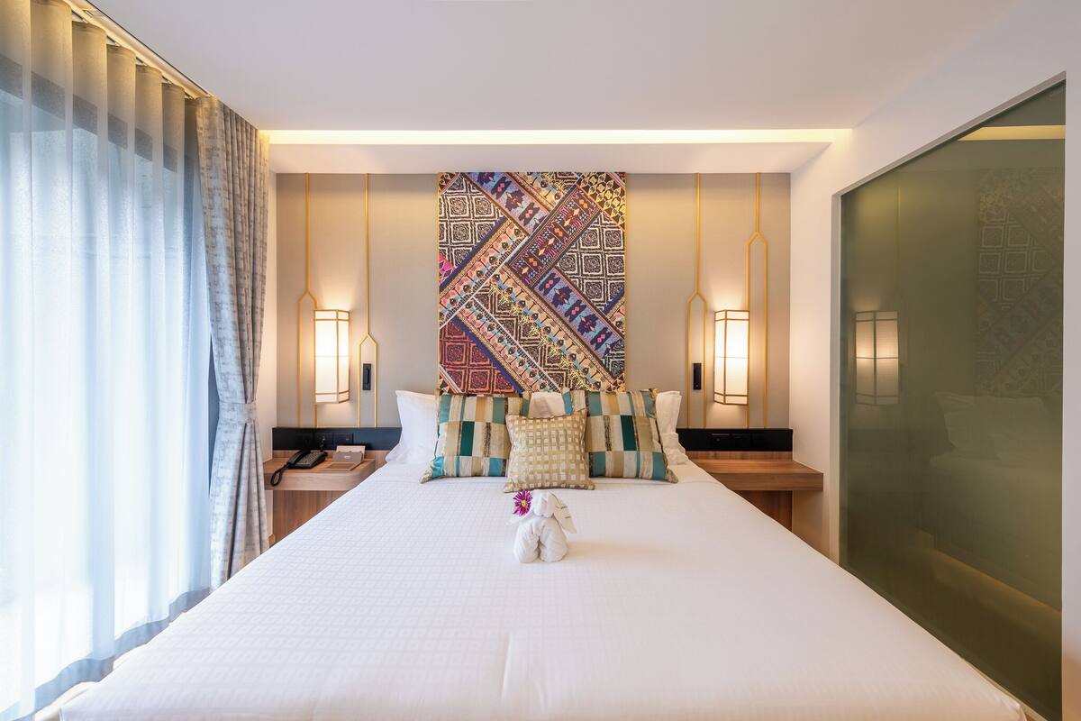 A hotel room with a large bed, two bedside tables, a patterned wall hanging, and a large window with curtains.