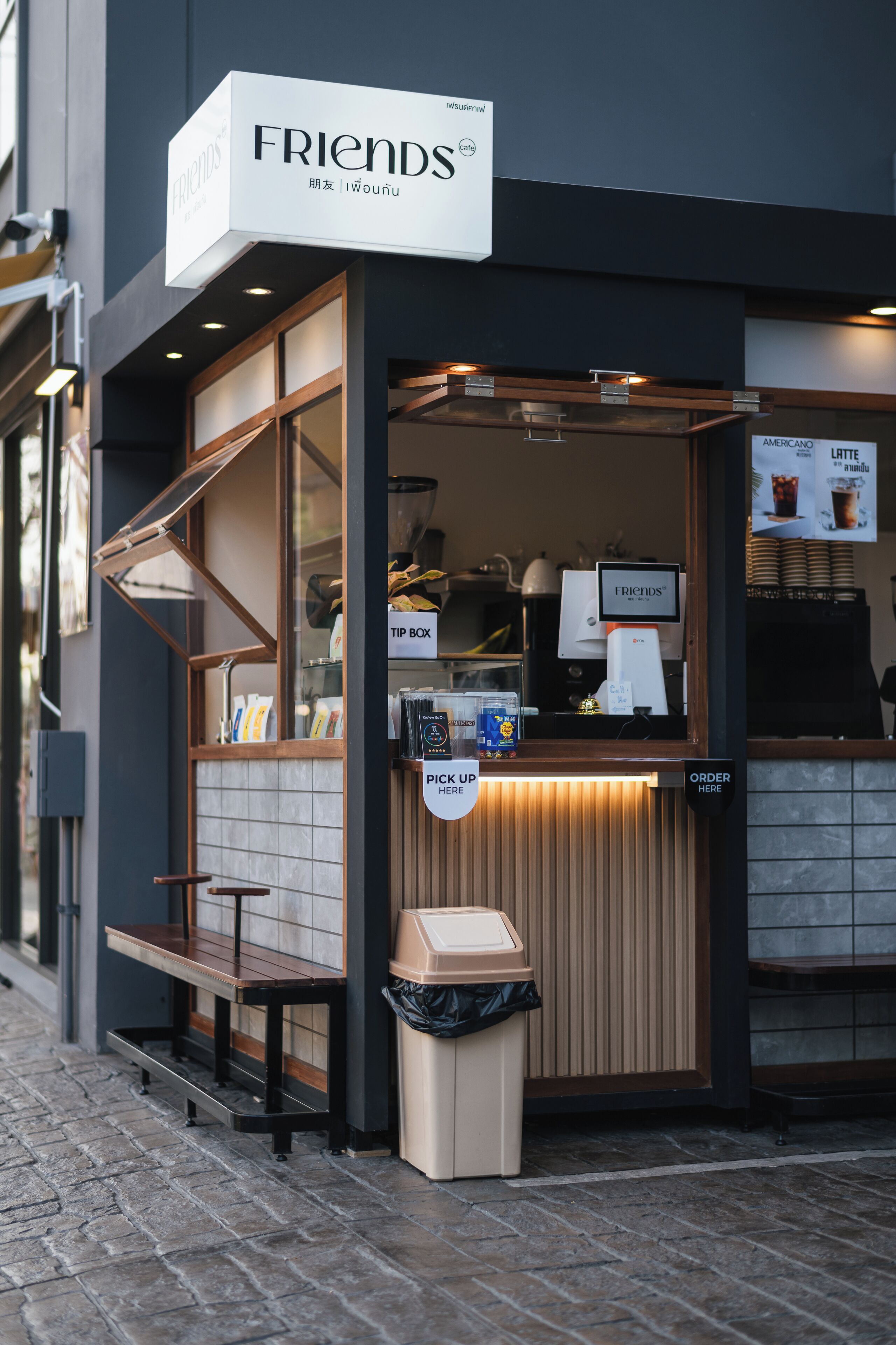 A Friends coffee shop with a pick-up window, a trash bin, and a menu board advertising Americano and Latte.
