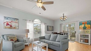 A living room with a grey sofa, a white coffee table, and a ceiling fan. There are two paintings on the wall, one of a beach scene and another of a dog. - Myrtlewood Resorts Serene Getaway Unit 105 (Myrtle Beach)