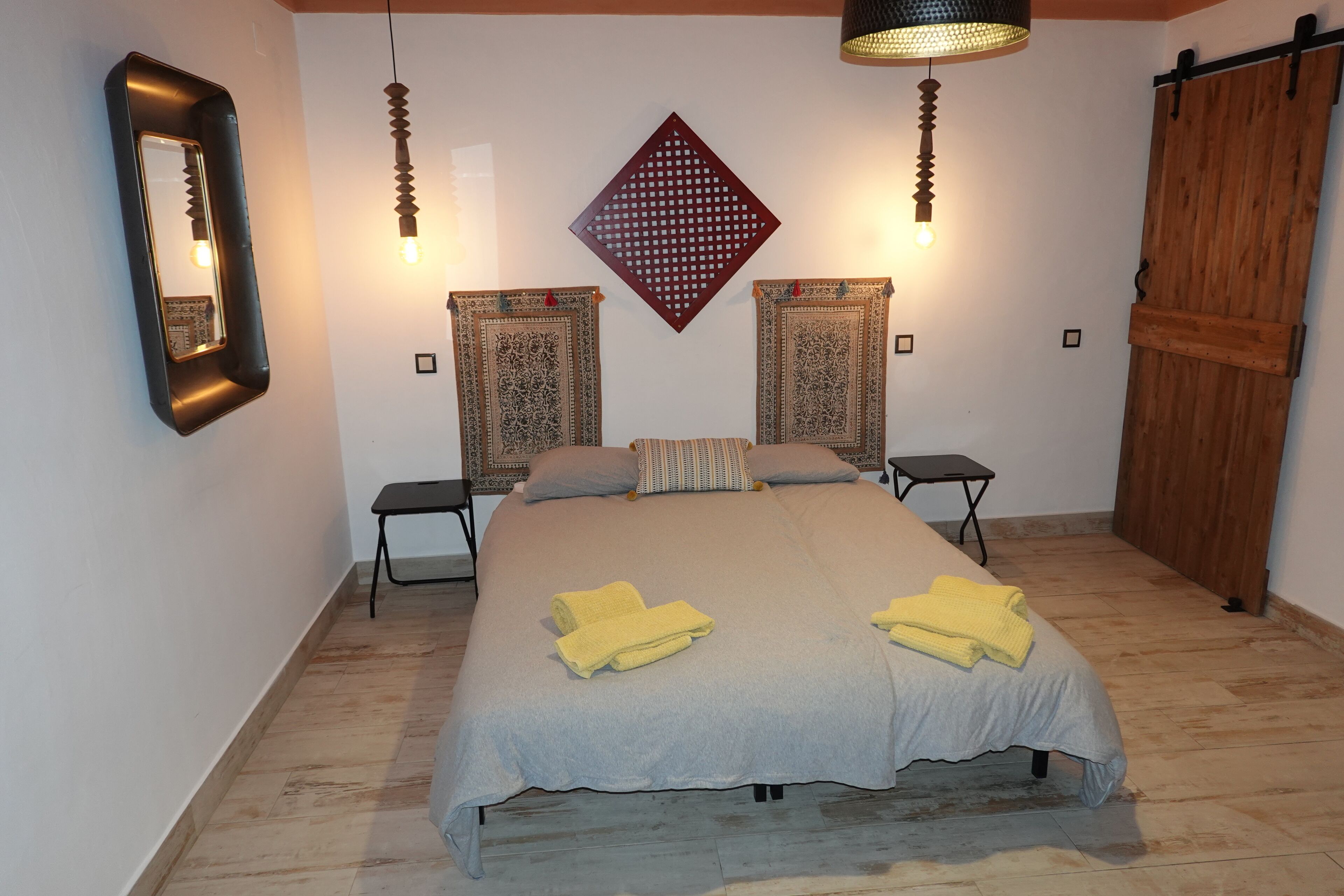 A bedroom with a bed, two nightstands, a mirror, patterned wall hangings, and a wooden wardrobe.