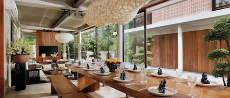 A modern dining area with a long wooden table set for a meal, surrounded by large windows and a view of greenery.