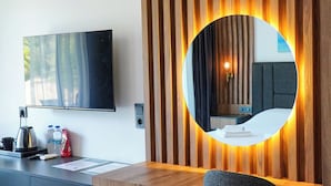 A modern hotel room with a wooden desk, a round mirror, a flat-screen TV, and a grey chair.