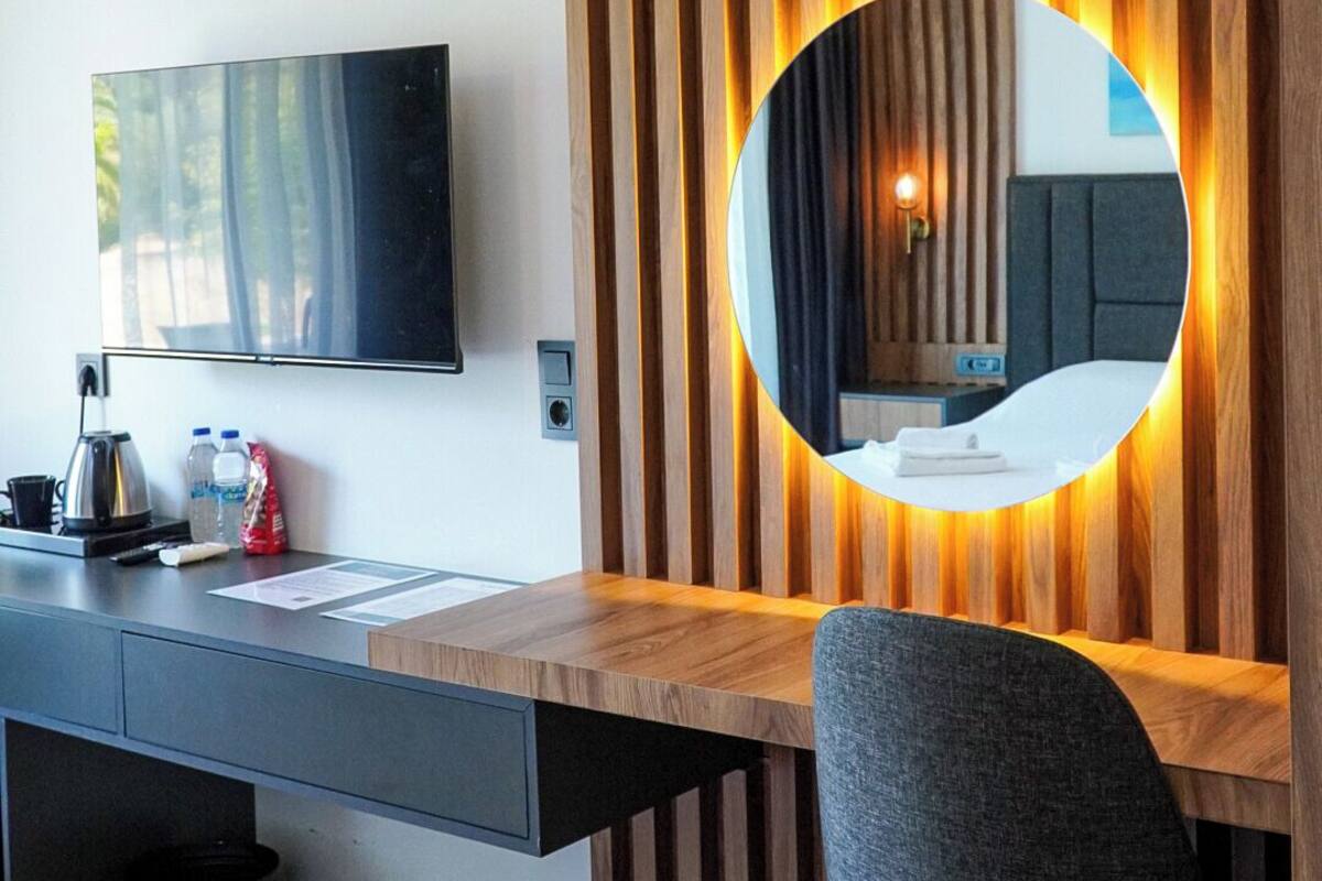 A modern hotel room with a wooden desk, a round mirror, a flat-screen TV, and a grey chair.
