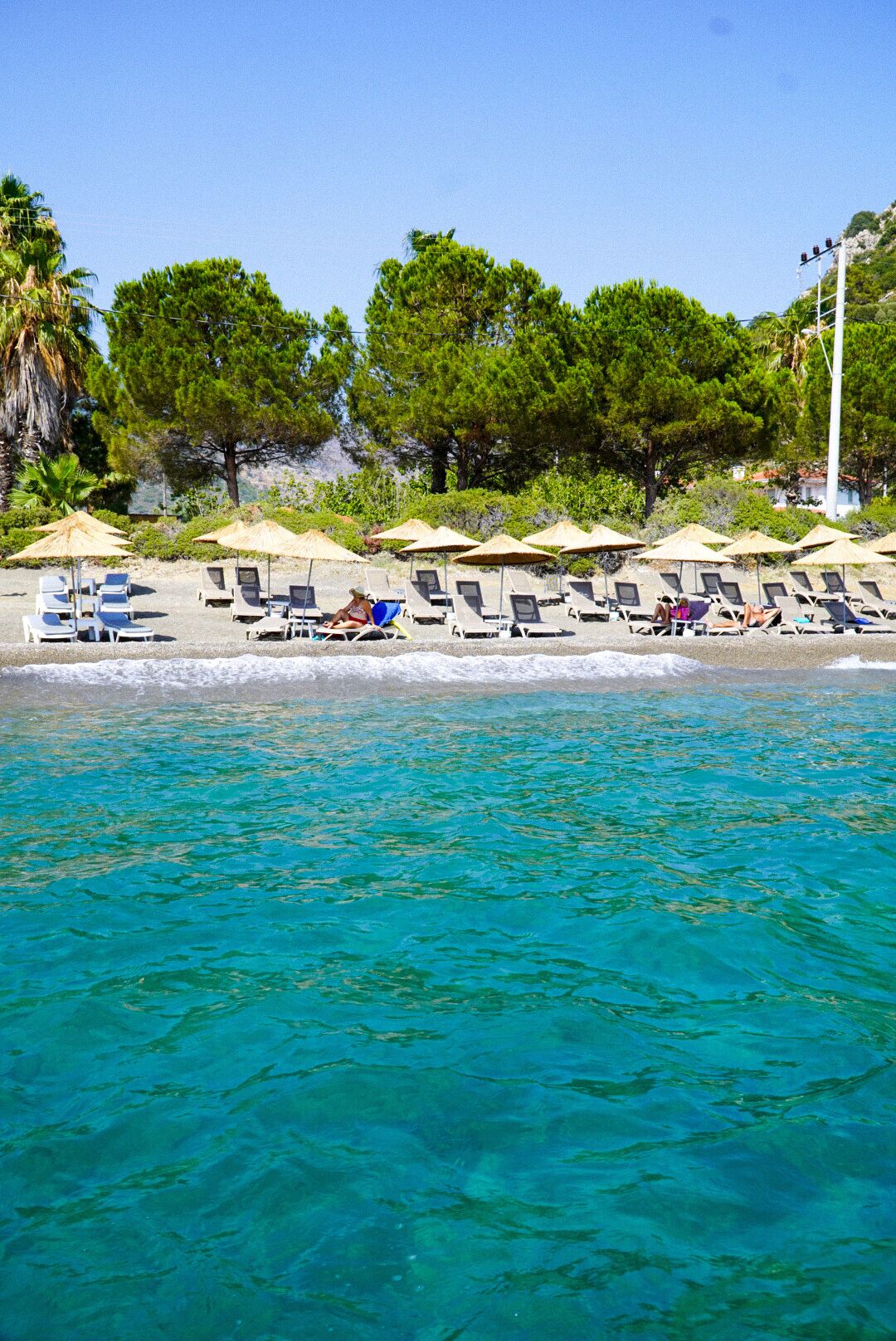 A beach with sun loungers and umbrellas, clear turquoise water, and trees in the background.
