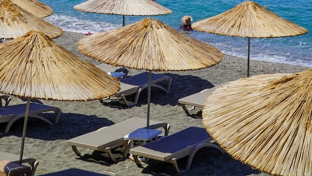 A beach with thatched umbrellas and lounge chairs, a rocky island in the background, and clear blue water.