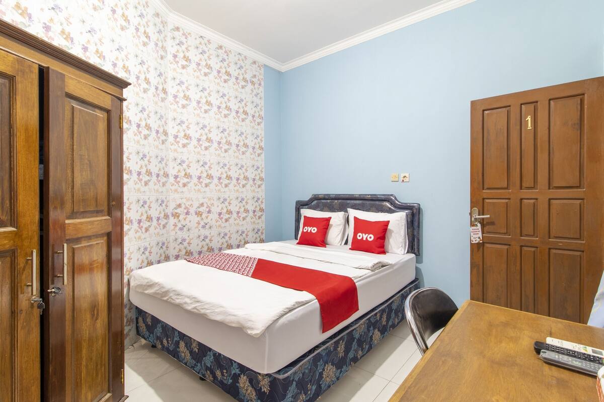 Een slaapkamer met een bed, een houten garderobe, een tafel met een stoel en een deur met een nummer.