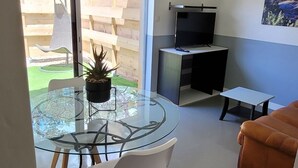 A modern living room with a glass dining table, white chairs, a brown sofa, a flat-screen TV, and a painting on the wall. - T2 with swimming pool in the heart of the pines (Saint-Julien)