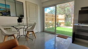 A modern living room with a glass dining table, white chairs, a sofa, a flat-screen TV, and a sliding glass door leading to a backyard with a wooden fence and greenery. - T2 with swimming pool in the heart of the pines (Saint-Julien)