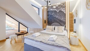 Ein modernes Schlafzimmer mit einem großen Bett, einem Dachfenster, einem Wandgemälde von Bergen und einem kleinen runden Tisch mit einer Lampe.