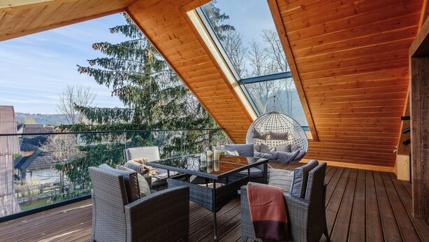 Eine Holzterrasse mit Glasdach, Korbmöbeln und Blick auf Bäume und Gebäude.