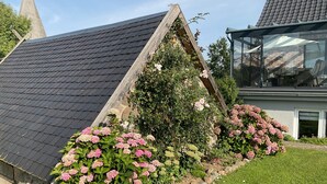 Een huis met een schuin dak, een tuin met roze hortensia's, en een aangrenzende kerk.