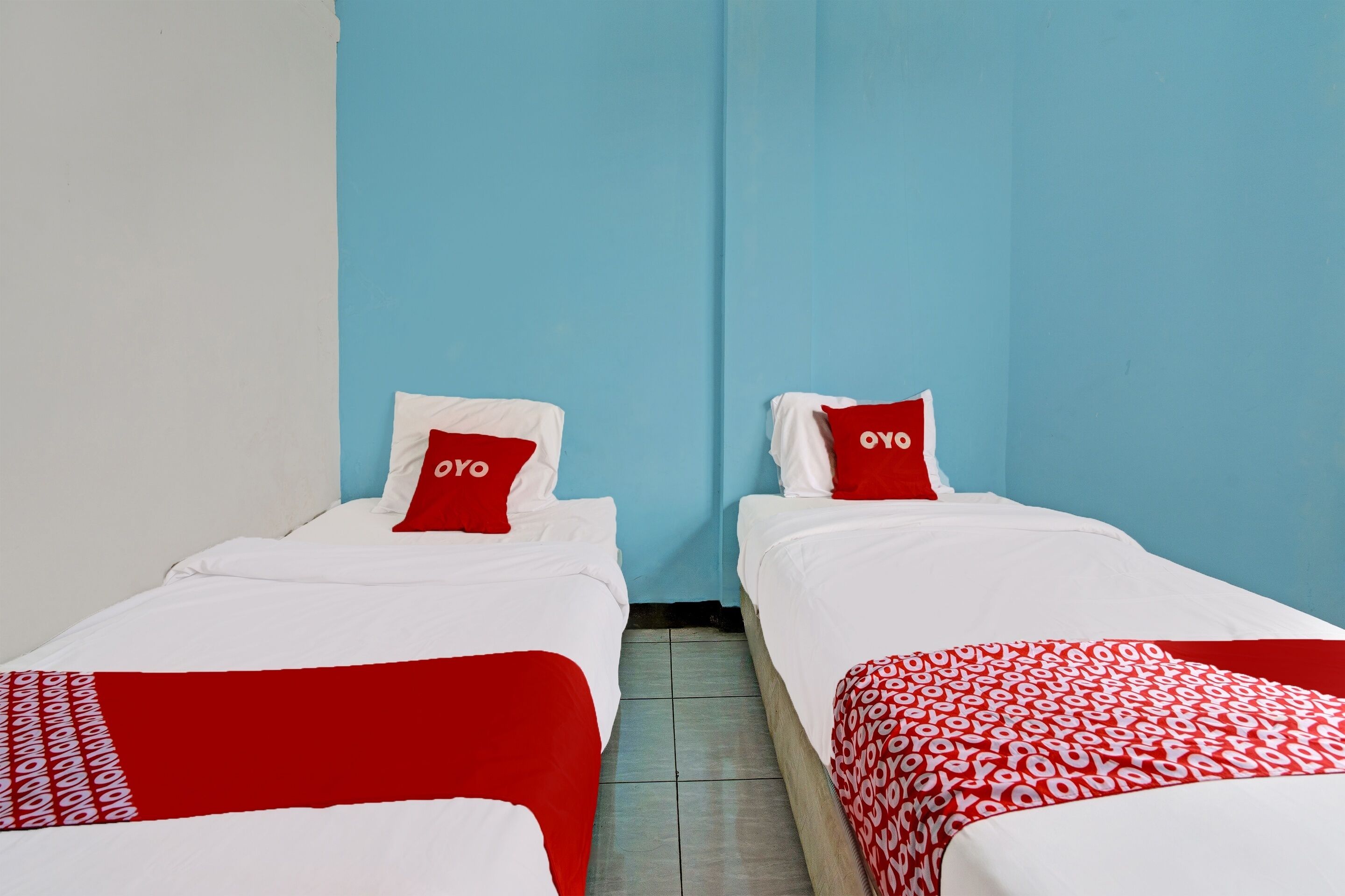 A hotel room with two single beds, each with a red and white bedspread and matching pillows. The room has light blue walls and a tiled floor.