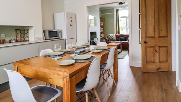 Un comedor con mesa de madera dispuesta para cuatro personas, sillas blancas, una cocina con electrodomésticos blancos y una puerta de madera.