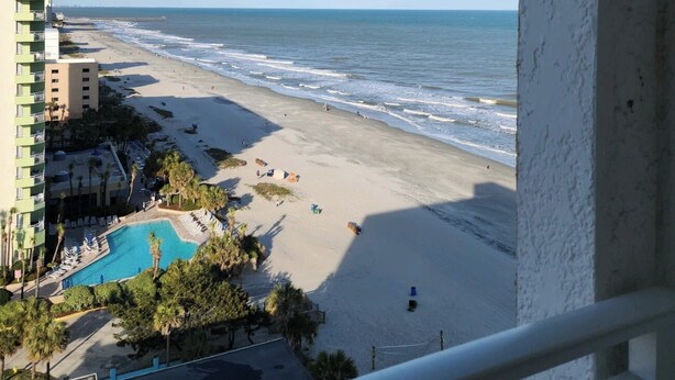 A beach view from a balcony overlooking a swimming pool and beachfront.