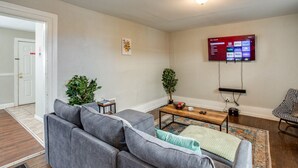 Una sala de estar con un sofá gris, una mesa de centro de madera, un televisor montado en la pared y una alfombra estampada.