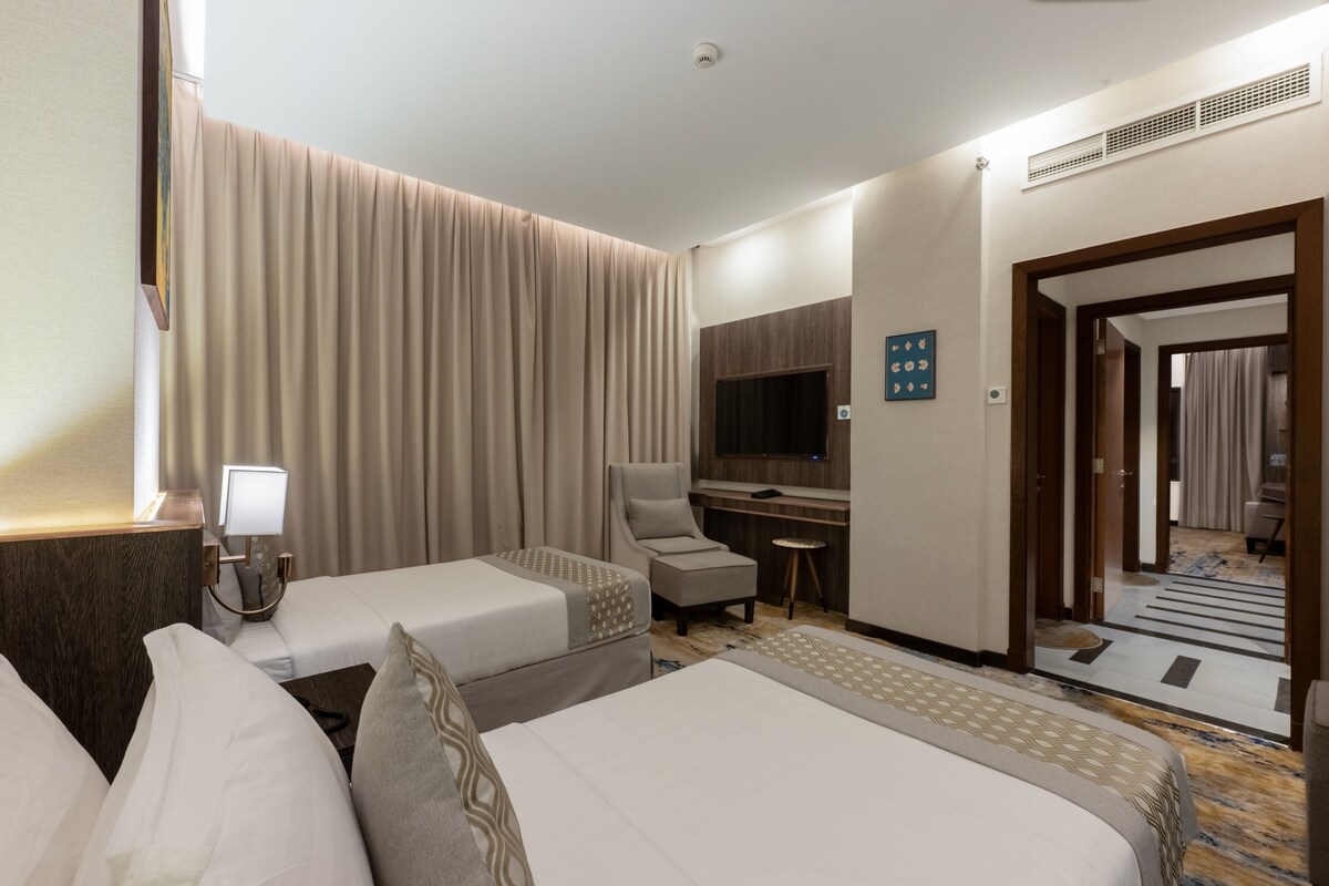 A hotel room with two beds, a desk, a chair, a TV, and a bathroom visible through an open door.