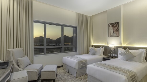A hotel room with two beds, a desk, a chair, and a large window with a mountain view. - Westin Mark Hotel (Madinah)