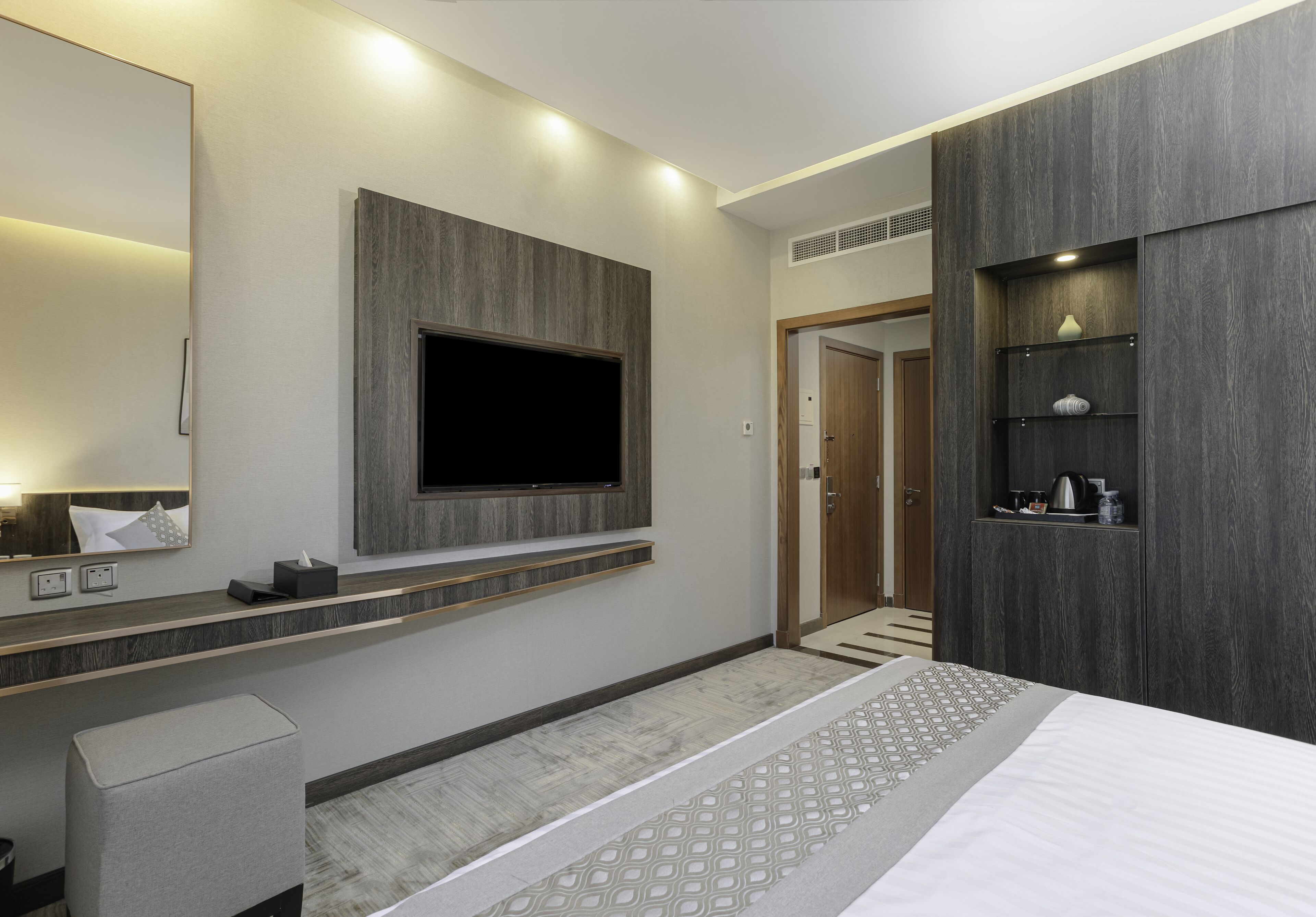 Una habitación de hotel moderna con una cama grande, un televisor montado, un tocador con espejo y un estante empotrado con artículos de aseo.