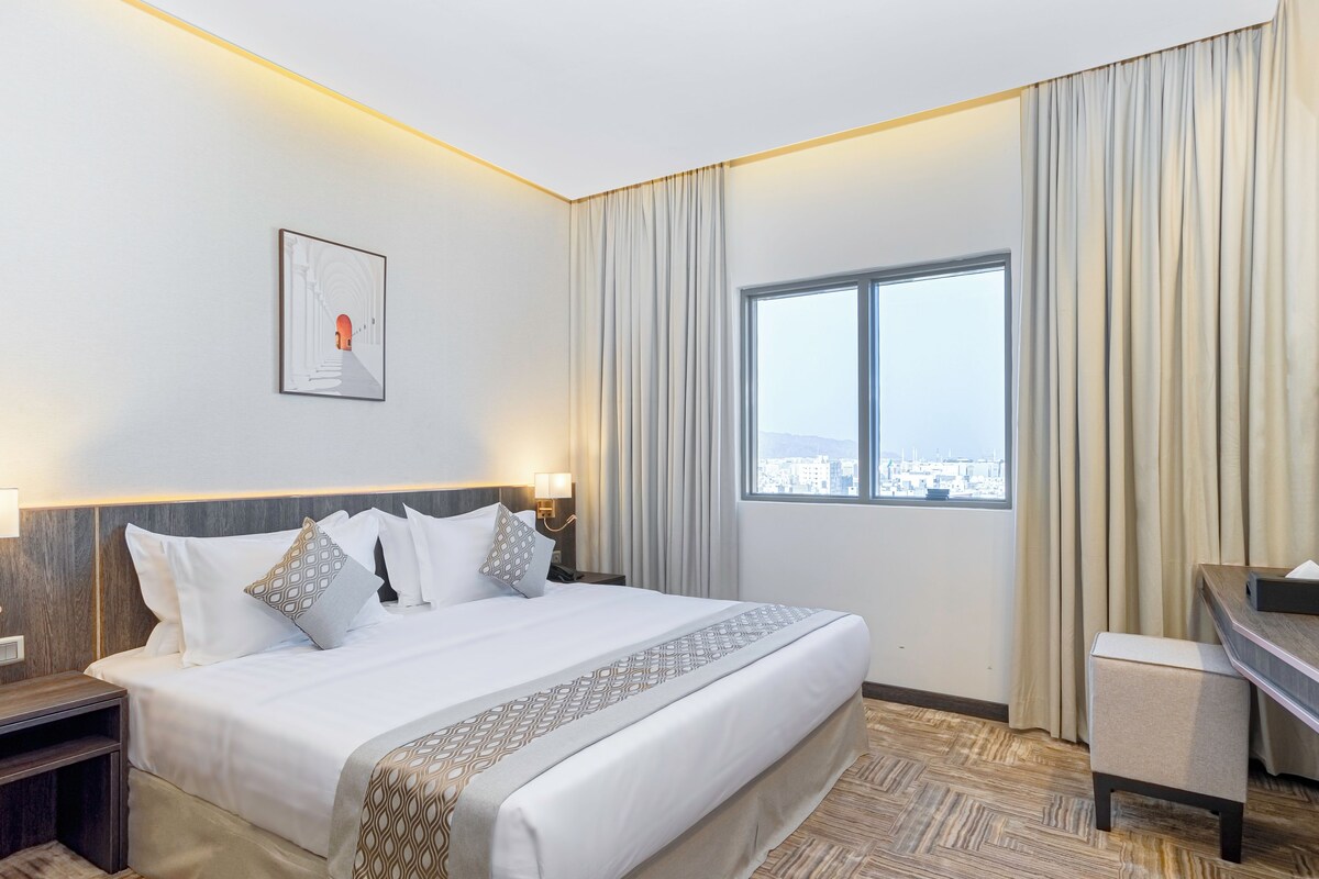 Ein Hotelzimmer mit einem großen Bett, einem Schreibtisch, einem Stuhl, einem Fenster mit Blick auf die Stadt und einem gerahmten Bild an der Wand.