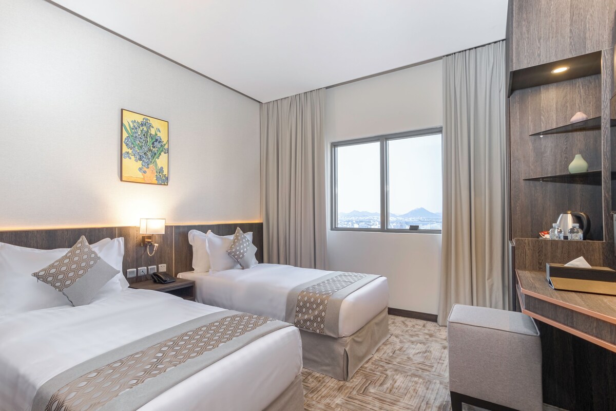 A hotel room with two beds, a desk, a nightstand, a lamp, a painting, and a window with a view.