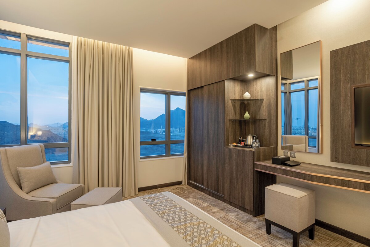 Habitación de hotel con cama, escritorio, silla, un espejo grande y vistas a montañas a través de las ventanas.
