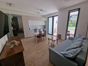 A modern living room with a grey sofa, wooden dining table, and chairs. There is a kitchen area with white cabinets and a large window.