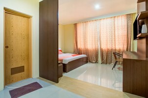 Habitación de hotel con puerta de madera, cama con colcha roja y blanca, escritorio con silla y un ventanal con cortinas.